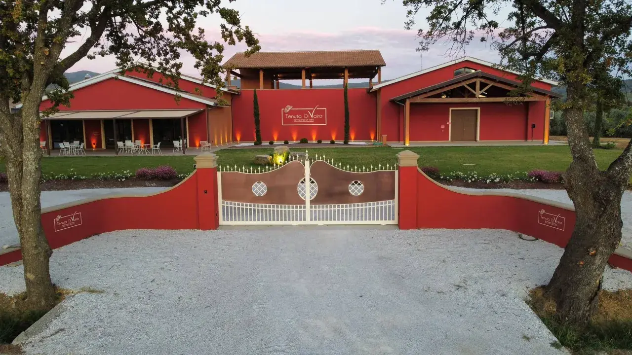 L'ingresso della Tenuta di Vaira a Bolgheri, con cancelli in rame e bianco, edifici rossi e un prato curato.