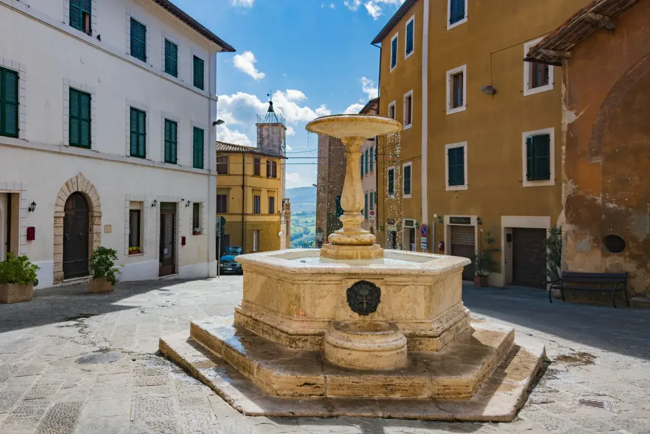 Fontana in pietra nel cuore dell'entroterra toscano, con palazzi storici e scorcio collinare.