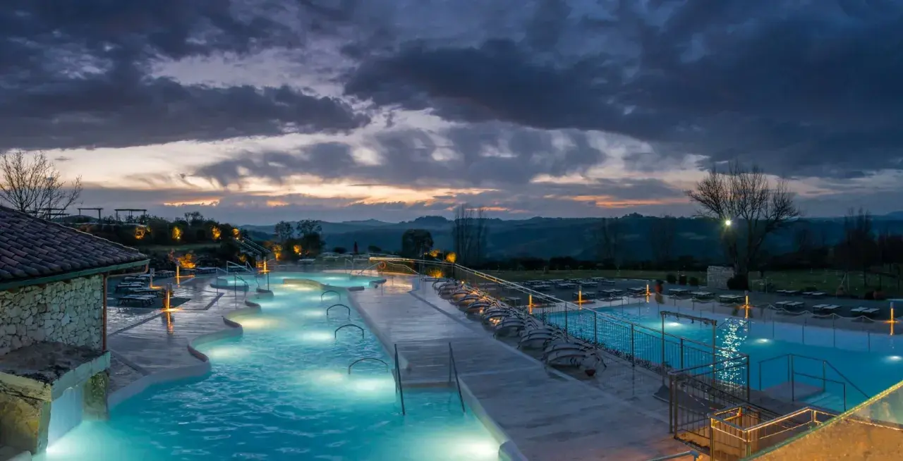 Piscine termali illuminate a Rapolano Terme al tramonto, con un cielo drammatico e colline toscane sullo sfondo.