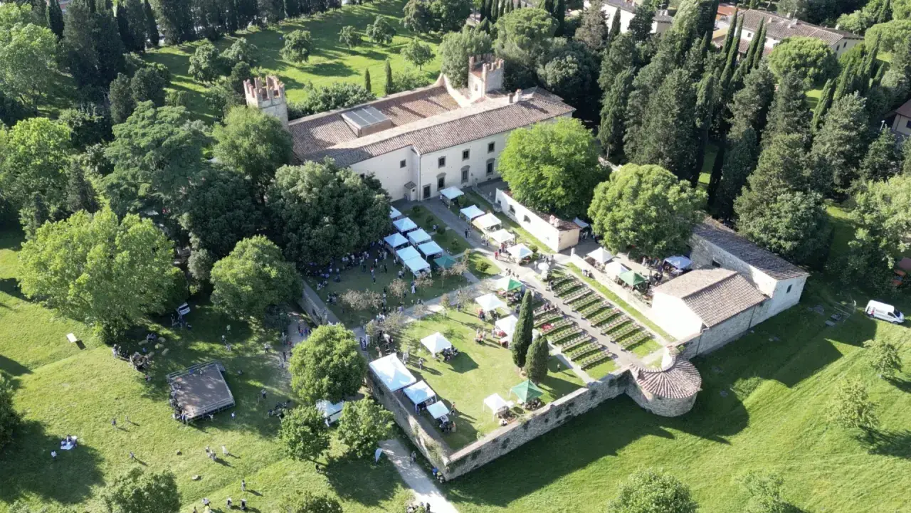 Mercato all'aperto nel parco dell'Acciaiolo, con bancarelle, persone e un edificio storico circondato da alberi.