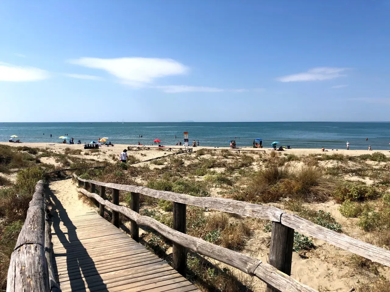 Passerella in legno conduce alla spiaggia di Tirrenia, con bagnanti e ombrelloni sotto il sole. Il mare calmo invita a un tuffo.