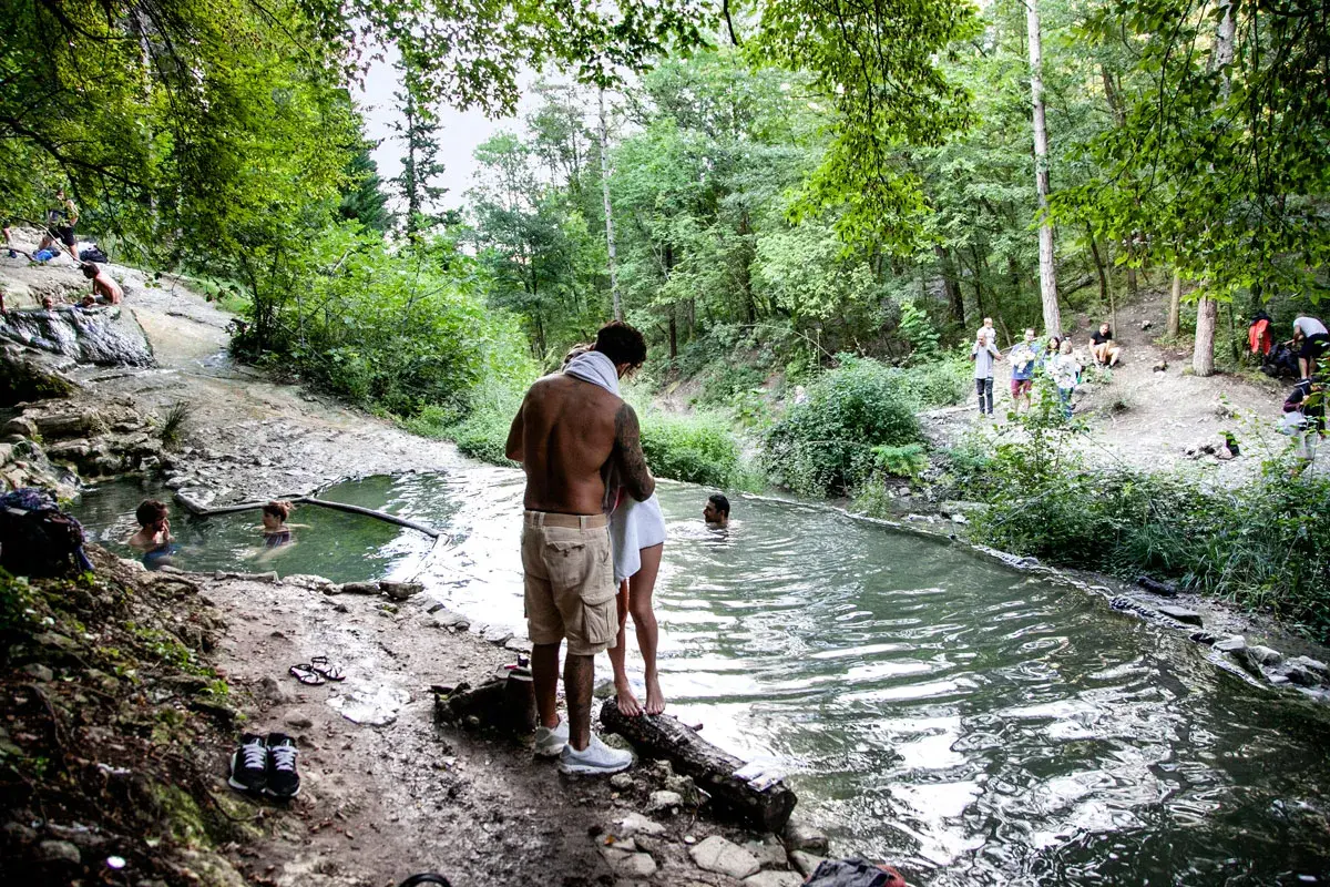 Persone si godono le acque termali ai Bagni San Filippo, un'oasi naturale in un bosco rigoglioso. Nonostante il nome, l'immagine evoca un'atmosfera estiva, non invernale.
