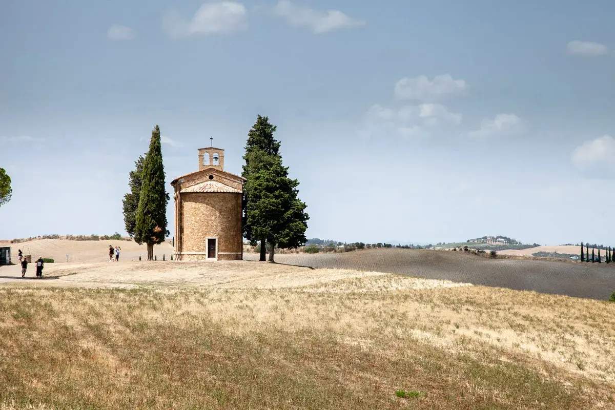 Iconica chiesetta Val d'Orcia, con cipressi e colline ondulate sotto un cielo nuvoloso.