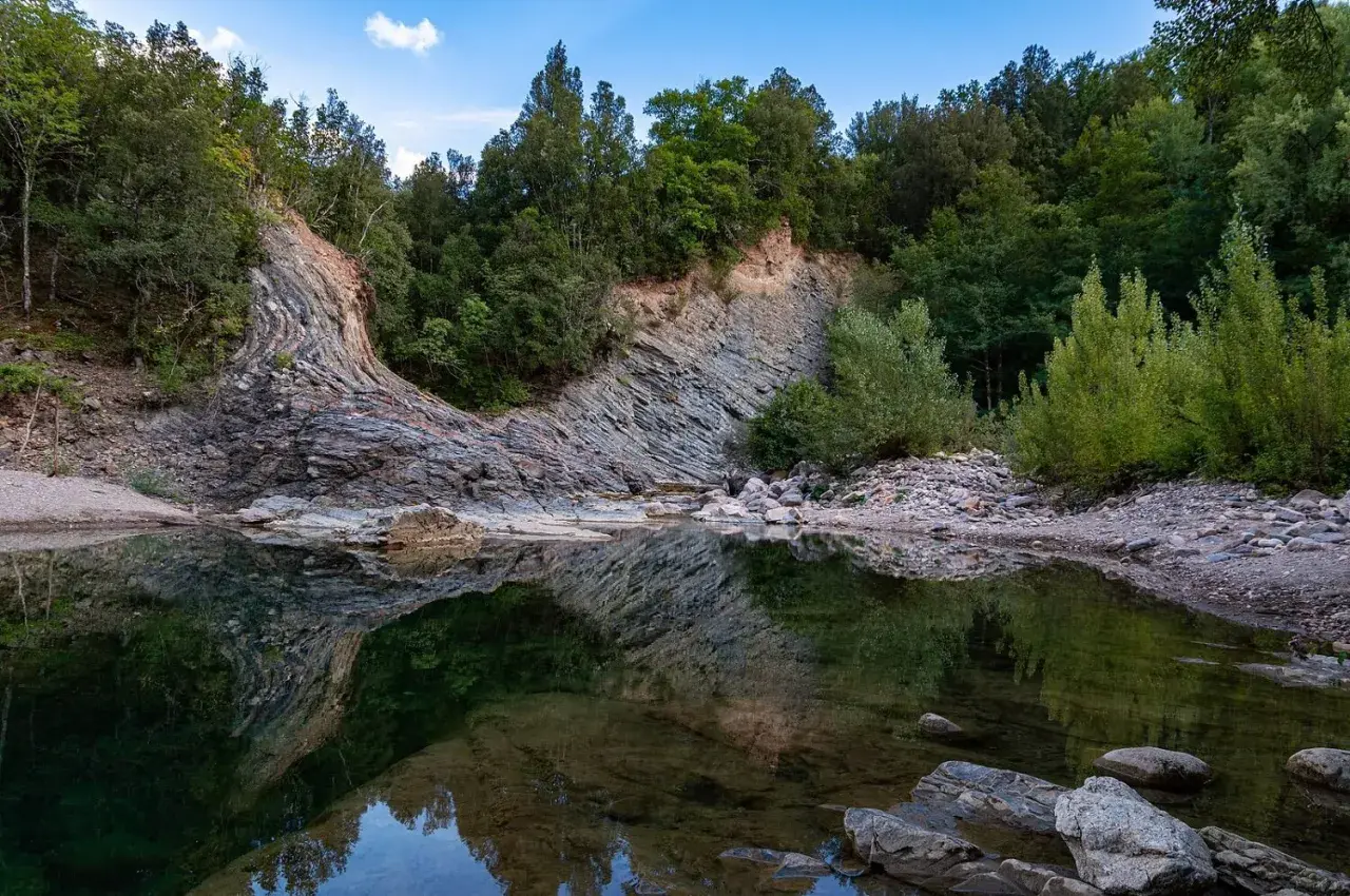Rocce stratificate e vegetazione lussureggiante si specchiano nelle acque tranquille del Val di Farma, un paesaggio naturale mozzafiato.