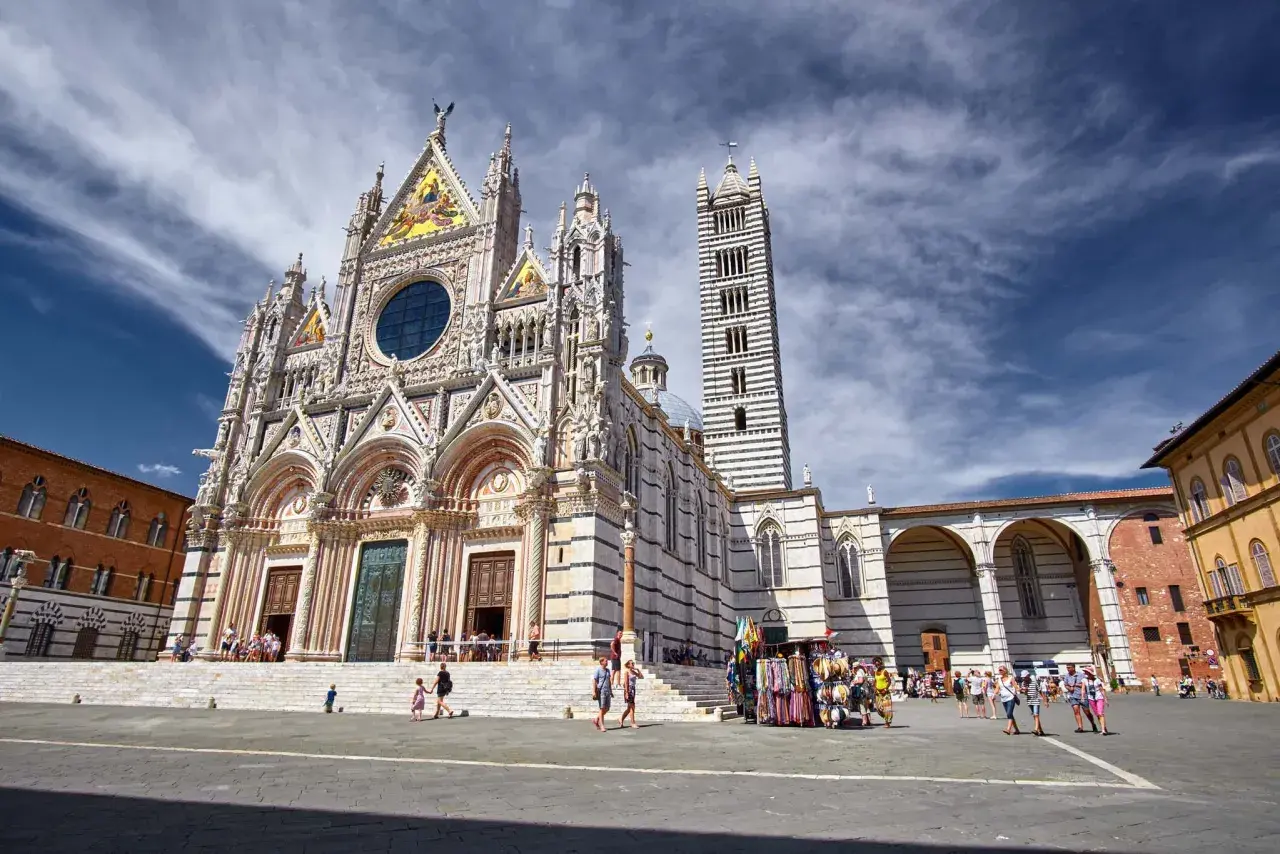 Magnifica facciata del Duomo di Siena, con la sua torre a strisce bianche e nere. Turisti passeggiano nella piazza antistante.