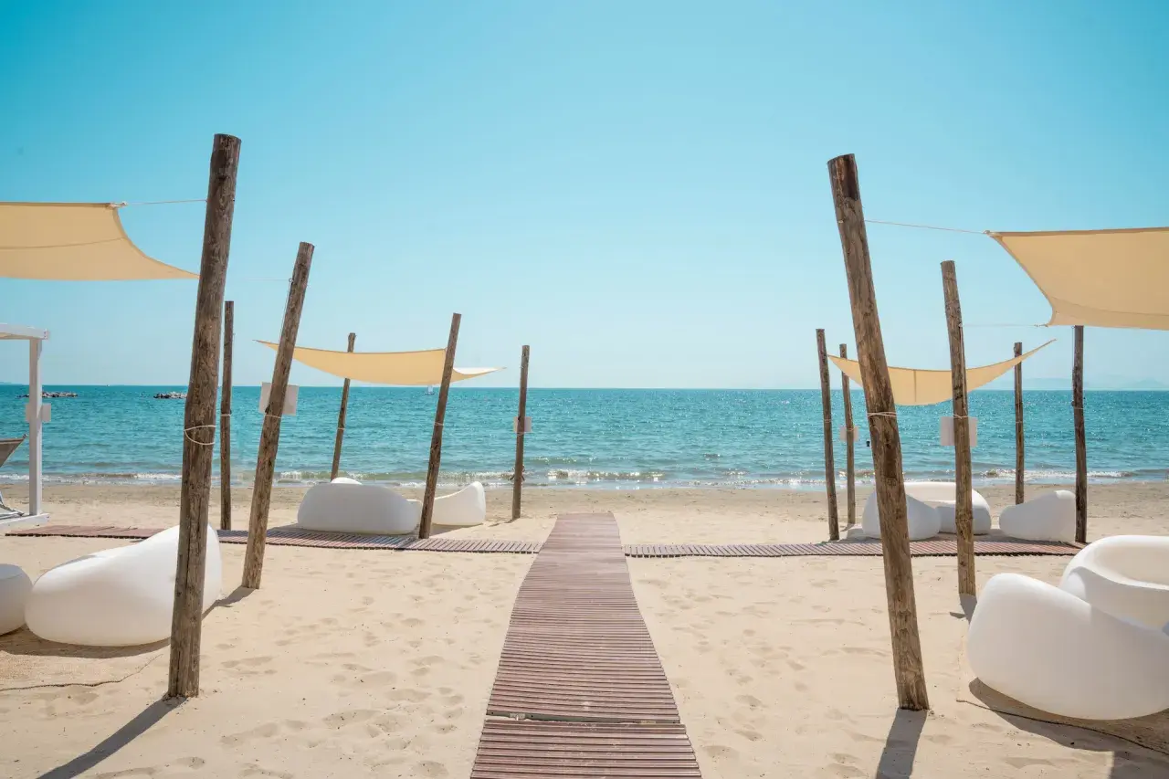Un percorso in legno conduce al **follonica mare**, dove lettini bianchi e tende color crema invitano al relax sotto il sole.