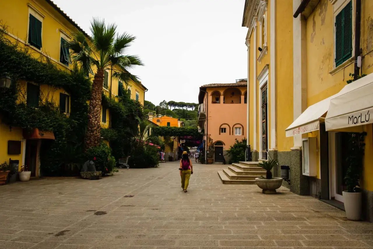 Una donna cammina in una piazza acciottolata del centro storico dell'Isola d'Elba, circondata da edifici color pastello e vegetazione lussureggiante.