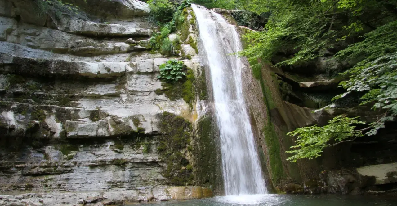 Cascata fragorosa su rocce stratificate, circondata da vegetazione lussureggiante. L'acqua cheta scende impetuosa, creando un velo bianco.