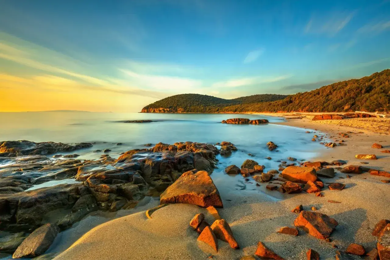 La Cala Martina al tramonto, con rocce scure e sabbia dorata bagnata da un mare calmo e cielo sfumato.