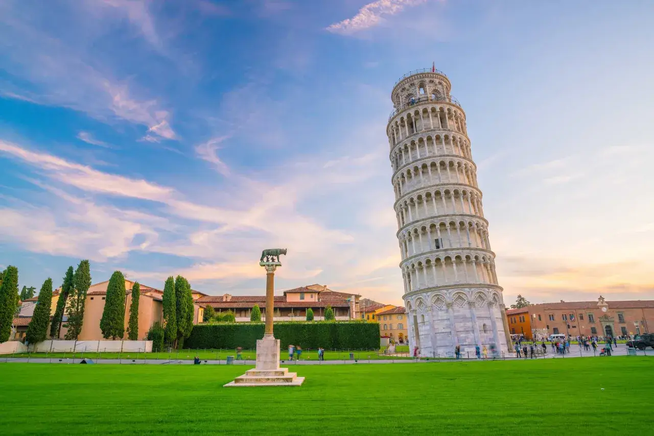 La celebre torre di Pisa, con la sua inconfondibile pendenza, si staglia contro un cielo azzurro e rosa. Un prato verde e un monumento completano la scena.