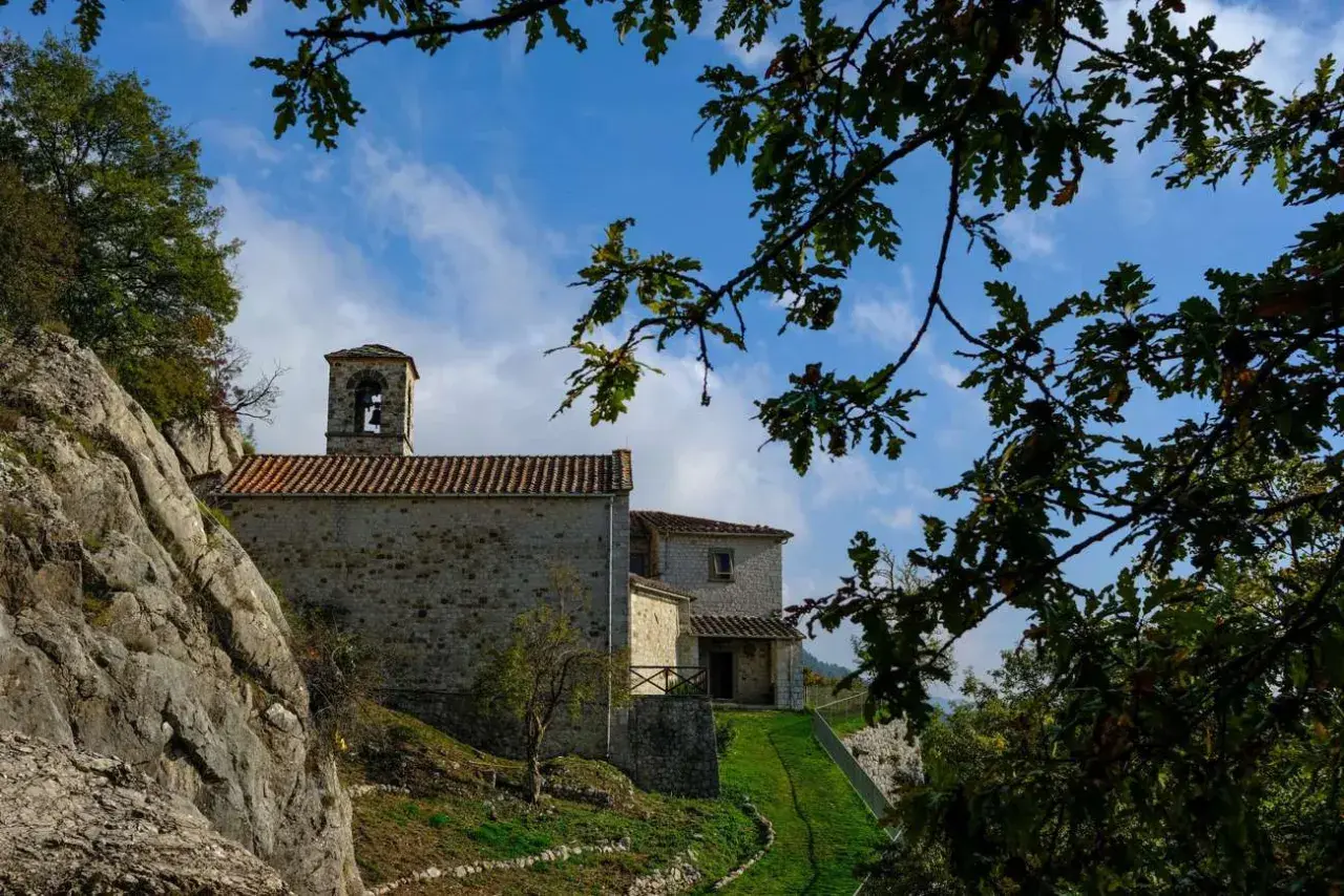 L'eremo di Cerbaiolo, con la sua pietra antica e il campanile, si staglia contro un cielo azzurro tra i rami di quercia.