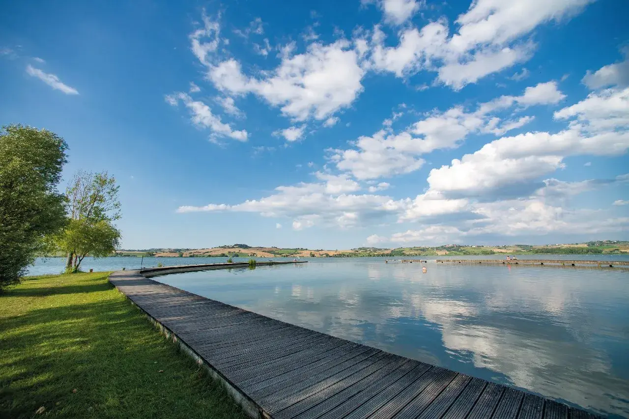 Passerella in legno su un lago tranquillo, con colline ondulate sullo sfondo. Un luogo idilliaco in Valdichiana, dove la natura incontra la serenità.