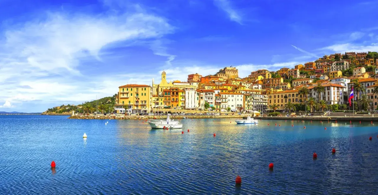 Vista del porto di Porto Santo Stefano, con case colorate che si arrampicano sulla collina e barche sull'acqua blu.
