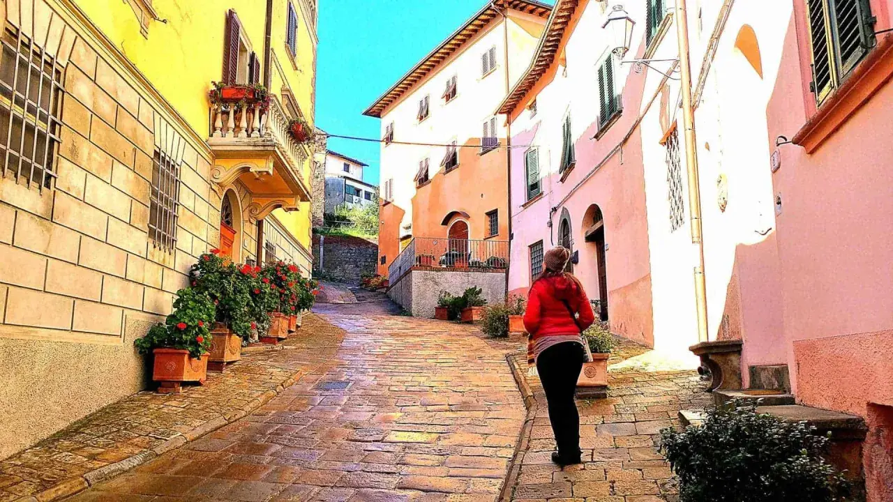 Una donna cammina su una strada acciottolata a Palaia, Pisa, tra case colorate e vasi di fiori rossi.