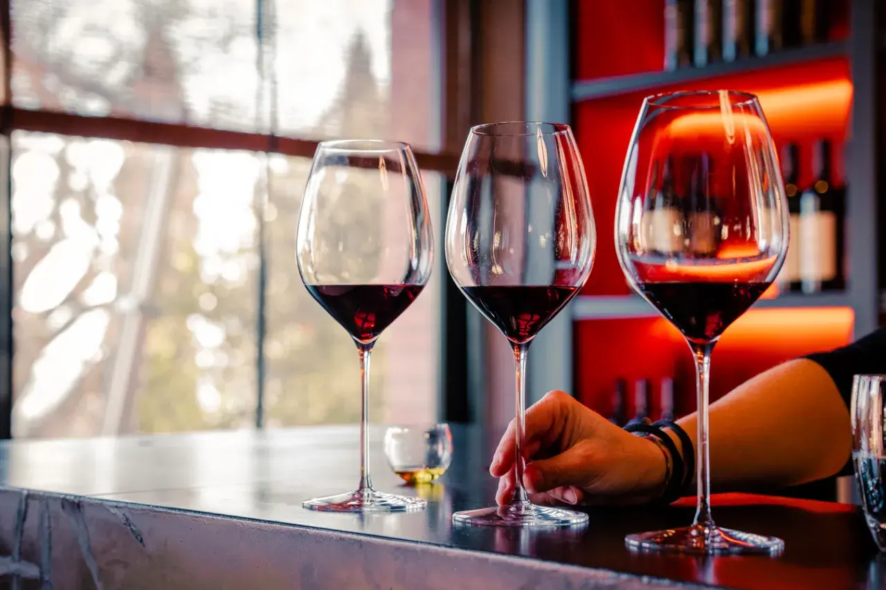 Calice di vino rosso su un bancone, con una mano che sfiora uno stelo. L'atmosfera è quella della cantina il poggio, con luci soffuse e bottiglie sullo sfondo.