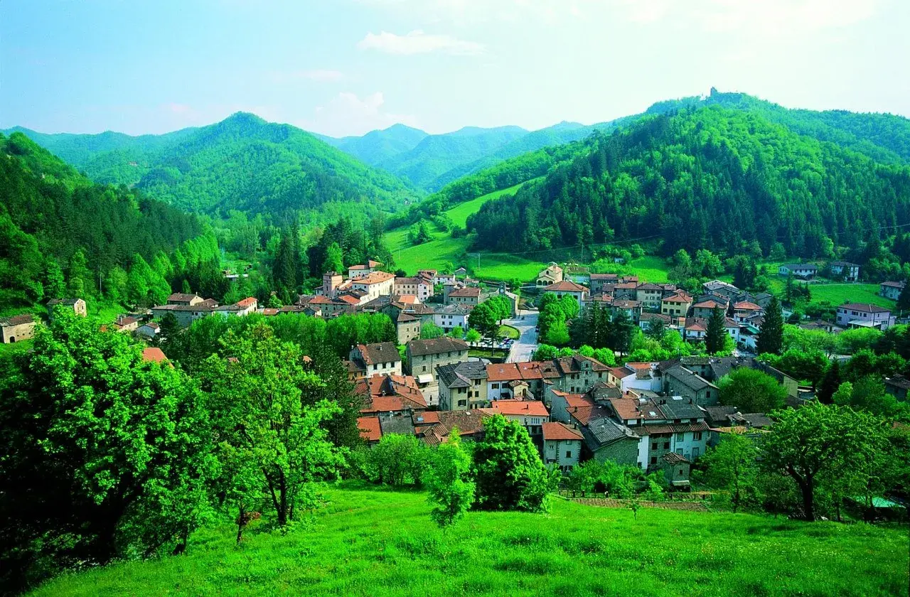 Un borgo incantevole, Palazzuolo sul Senio, immerso nel verde lussureggiante delle colline appenniniche. Case con tetti rossi si aggrappano ai pendii, circondate da alberi rigogliosi e un cielo azzurro.
