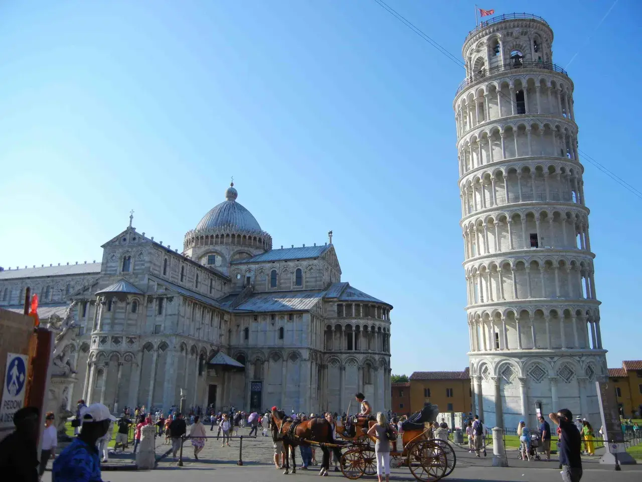 Visitare Pisa: la Torre Pendente e il Duomo svettano sotto un cielo azzurro, mentre turisti e una carrozza trainata da cavalli animano la piazza.