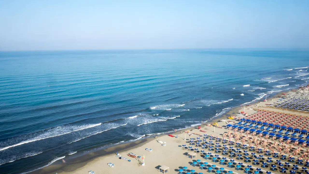 Ampia spiaggia attrezzata al **Lido di Camaiore**, con file ordinate di ombrelloni blu e arancioni che si estendono verso il mare calmo e azzurro.