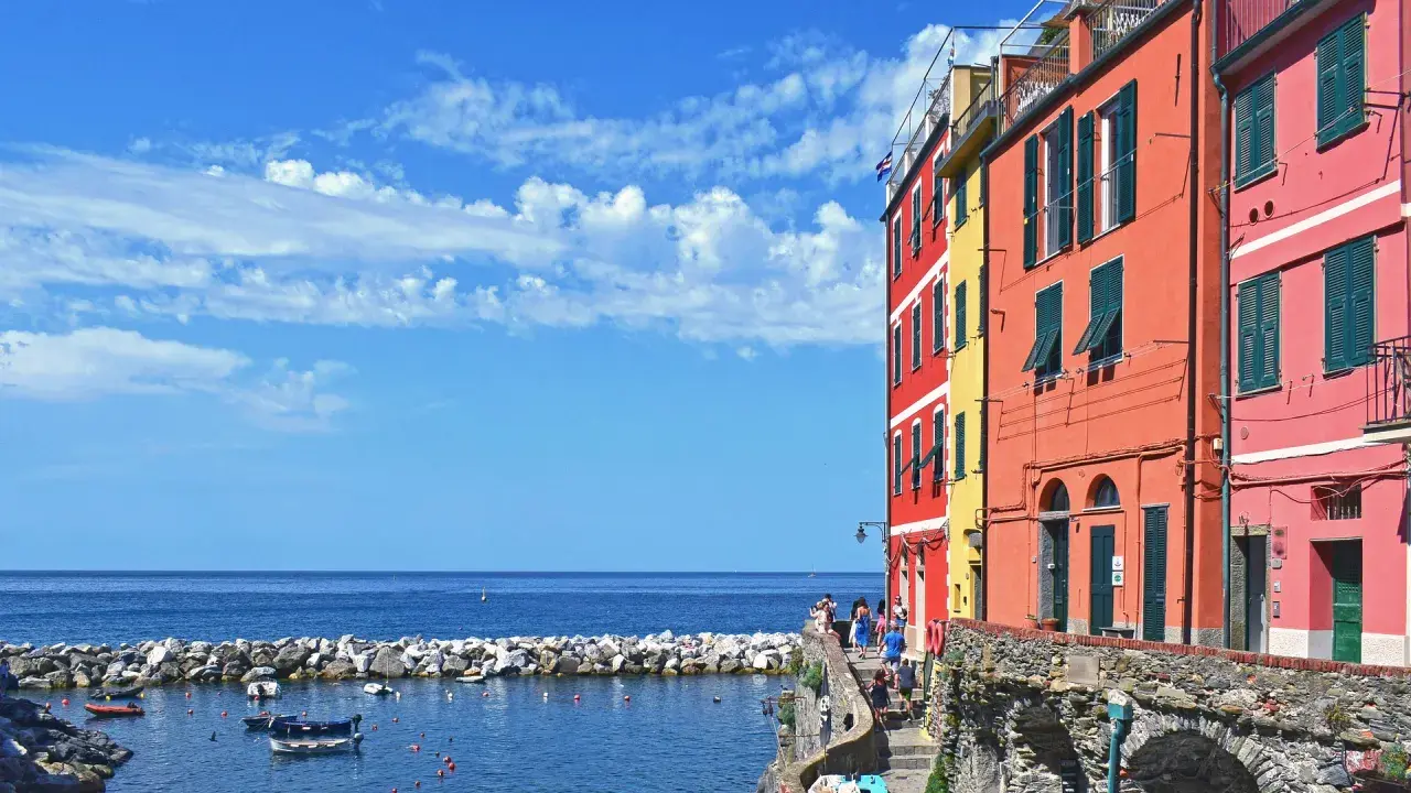 Case, a Firenze c'è il mare! Case colorate affacciate su un porto con barche, sotto un cielo azzurro con nuvole bianche.