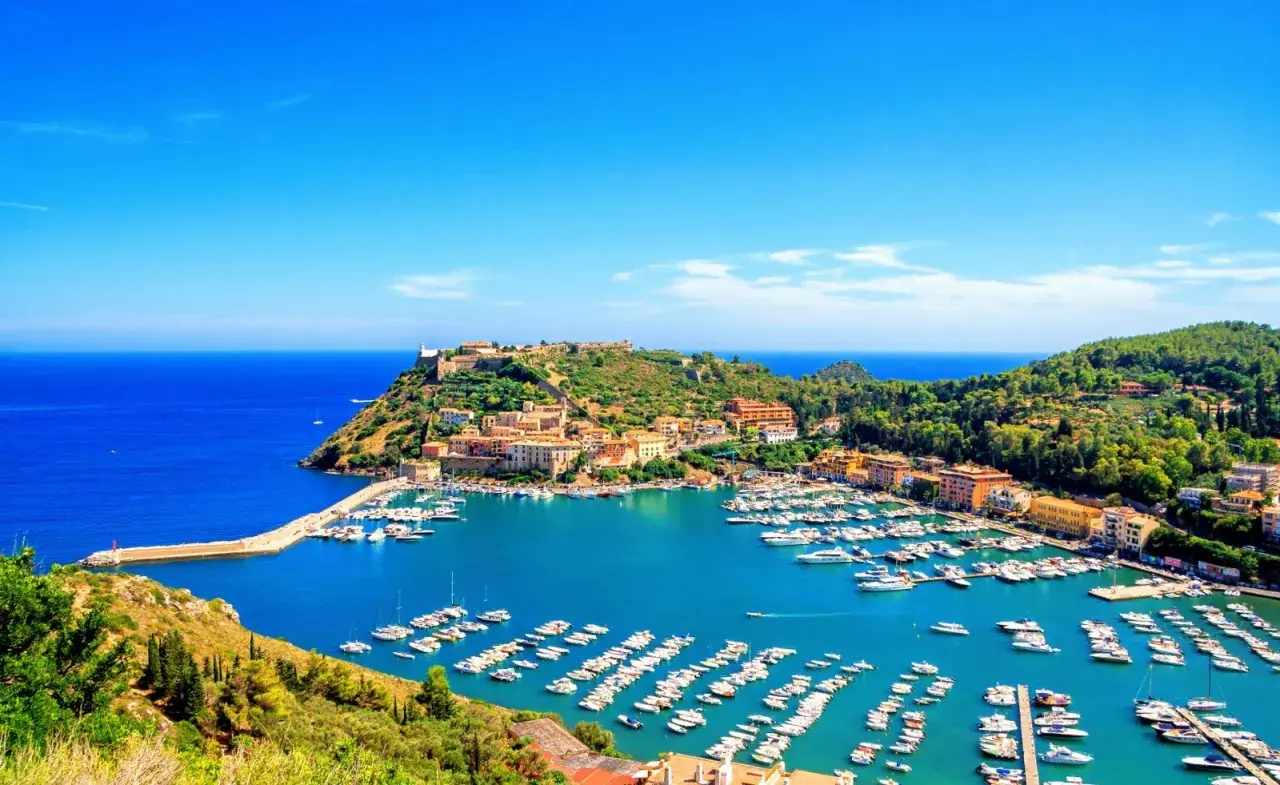 Porto Ercole cosa vedere: un incantevole porto con barche, case colorate e un castello su una collina verde, bagnato da un mare azzurro.