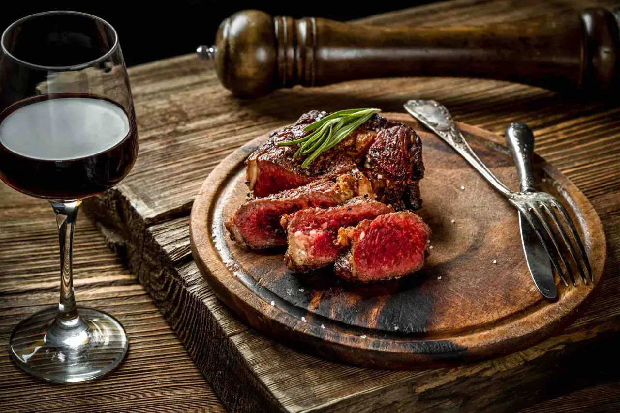Bistecca succulenta con rosmarino, accompagnata da un bicchiere di vino tannico e posate d'argento su un tagliere di legno rustico.