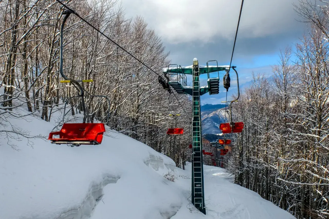 Seggiovie rosse e innevate salgono verso il cielo azzurro, attraversando un bosco spoglio. Un paesaggio invernale che ricorda il **centro** di **Abetone**.