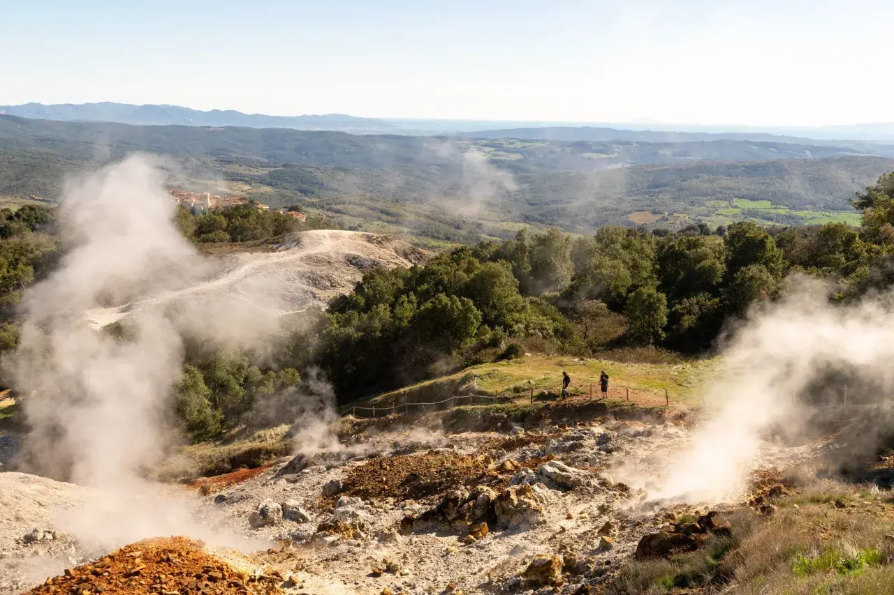 Vapori emergono dal terreno in un paesaggio collinare, con un villaggio in lontananza. Un geyser in Italia che incanta i visitatori.