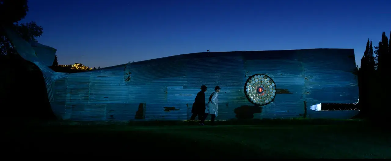 Due uomini osservano un'installazione artistica a forma di balena nel giardino, un viaggio di ritorno verso la luce.