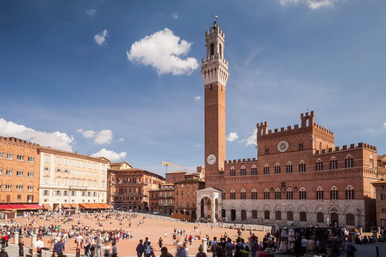 La Piazza del Campo di Siena, con la Torre del Mangia svettante, brulica di gente sotto un cielo azzurro.