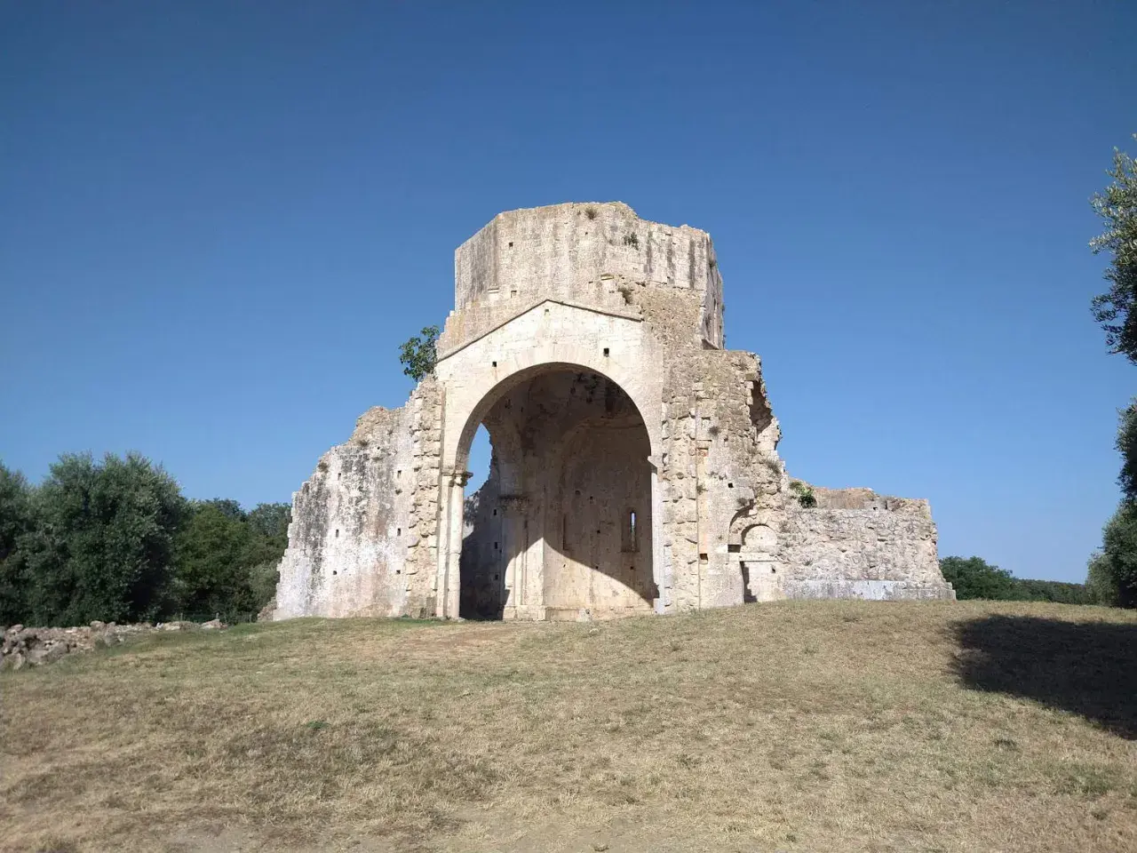 Rovine dell'abbazia di San Bruzio sotto un cielo azzurro, con alberi sullo sfondo.