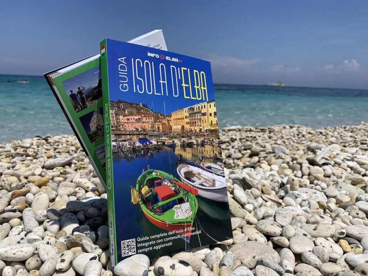 Guida turistica "Isola d'Elba" su una spiaggia di ciottoli, con il mare azzurro sullo sfondo. Perfetta per pianificare la tua vacanza.