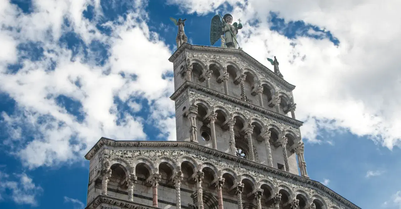 La facciata riccamente decorata della chiesa San Michele a Lucca, con un angelo in cima, si staglia contro un cielo azzurro e nuvoloso.