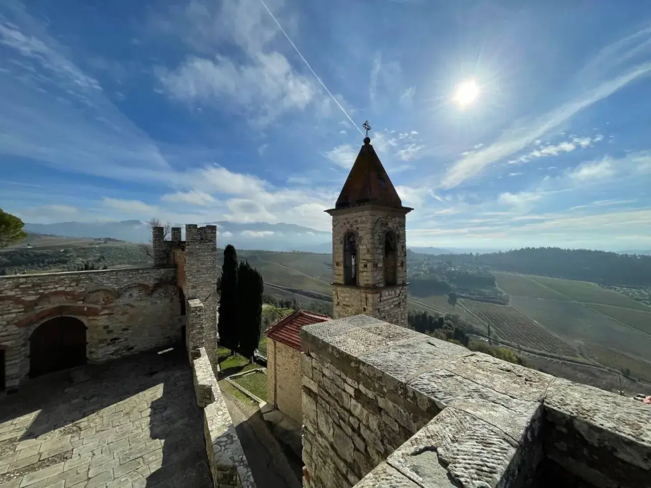Torre campanaria e mura antiche del castello Frescobaldi, con vigneti e colline toscane sotto un cielo azzurro e soleggiato.