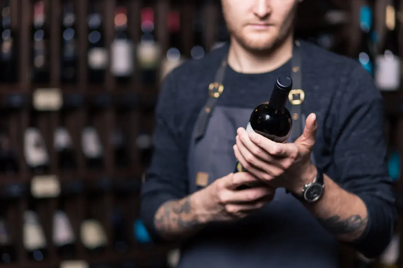 Un sommelier mostra una bottiglia di **vini toscani rossi pregiati**, con scaffali pieni di bottiglie sullo sfondo.