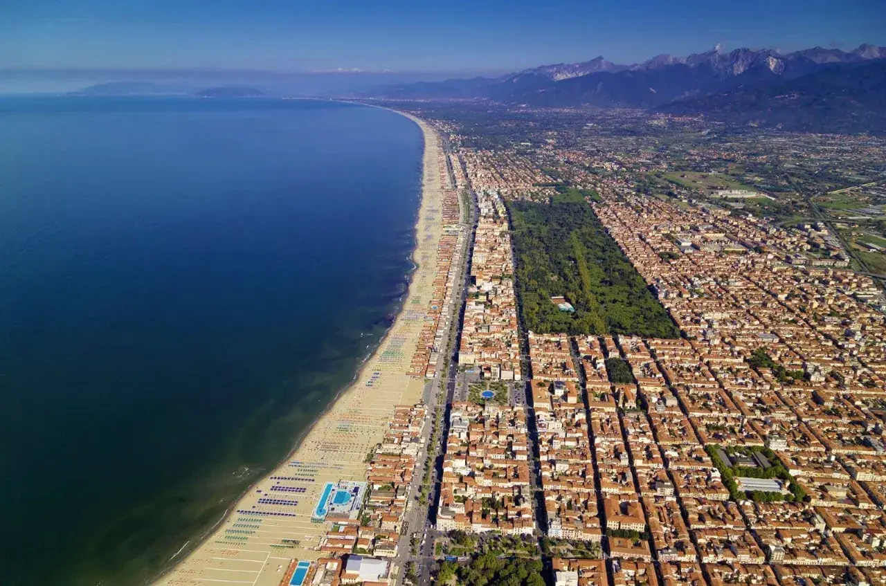 Vista aerea della costa di Massa Carrara, con il mare blu intenso, la lunga spiaggia attrezzata e la città che si estende verso le montagne.