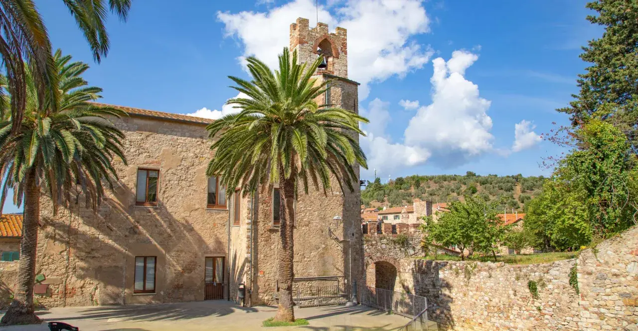 Un antico campanile in pietra a Suvereto, Livorno, svetta tra palme rigogliose e un cielo azzurro con nuvole bianche.