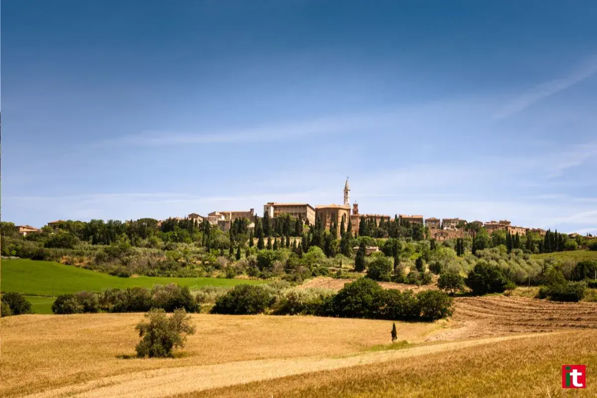 Un borgo medievale domina la Val d'Orcia, circondato da campi dorati e cipressi sotto un cielo azzurro.