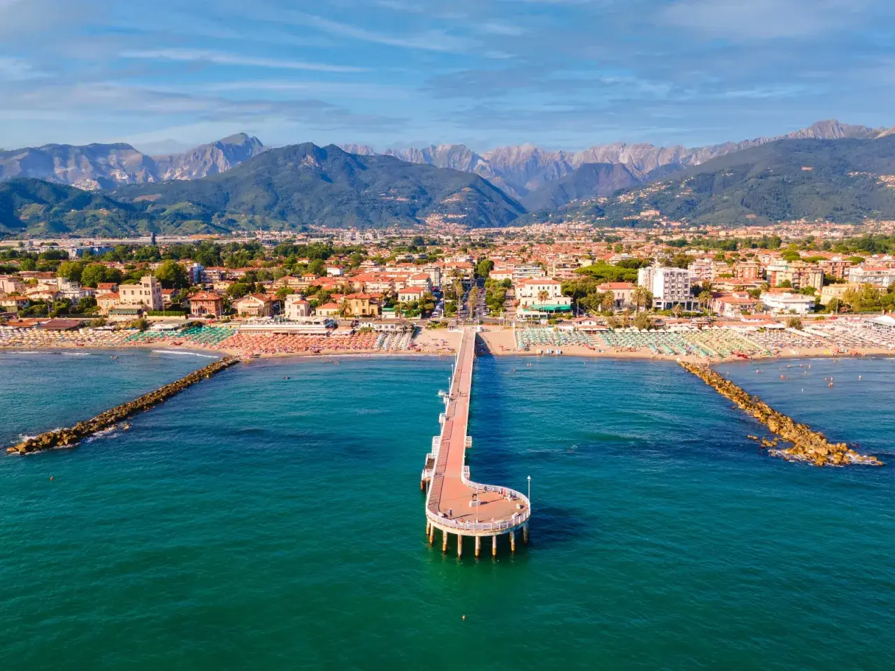 Un molo si estende nel mare turchese di Marina di Massa, con spiagge affollate e montagne sullo sfondo.