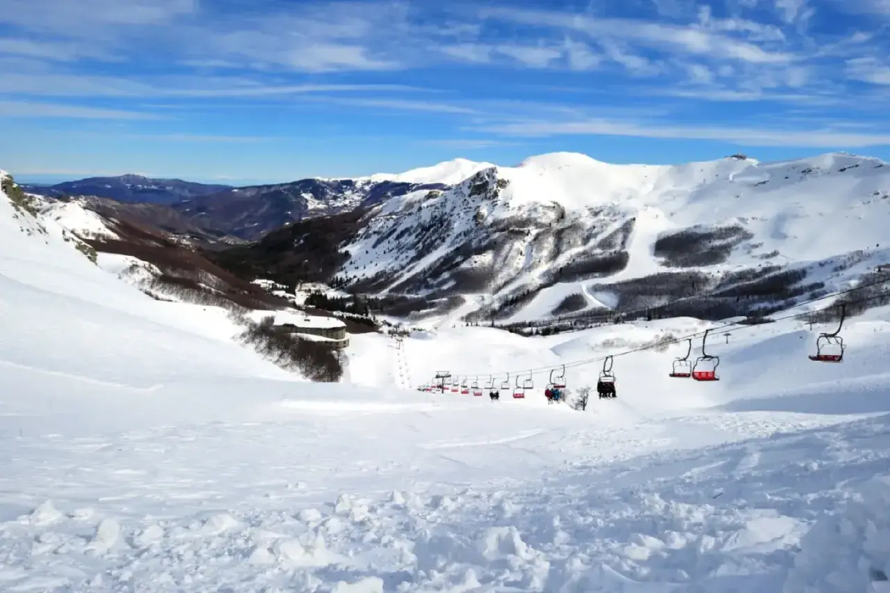 Impianti di risalita rossi trasportano sciatori su piste innevate ad Abetone, con montagne sullo sfondo sotto un cielo azzurro.