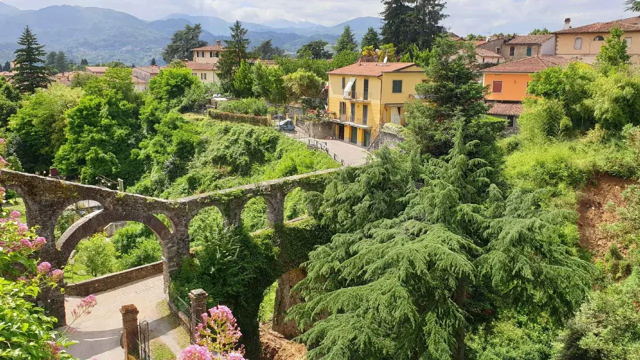 Un antico acquedotto in pietra, ricoperto di vegetazione, domina il paesaggio collinare dove si trova Barga. Case gialle e arancioni si stagliano tra il verde lussureggiante.