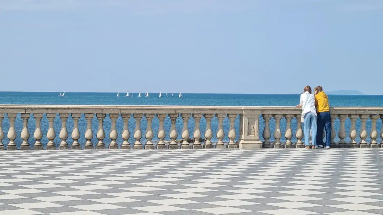 Due persone osservano le barche a vela sul mare, con la vista che ricorda la bellezza del lungomare di Livorno, vicino all'acquario.