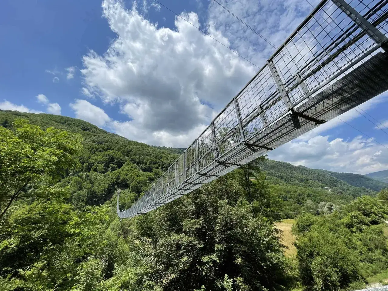 Il ponte sospeso delle Ferriere si estende su una valle verdeggiante, con un cielo azzurro e nuvole bianche.