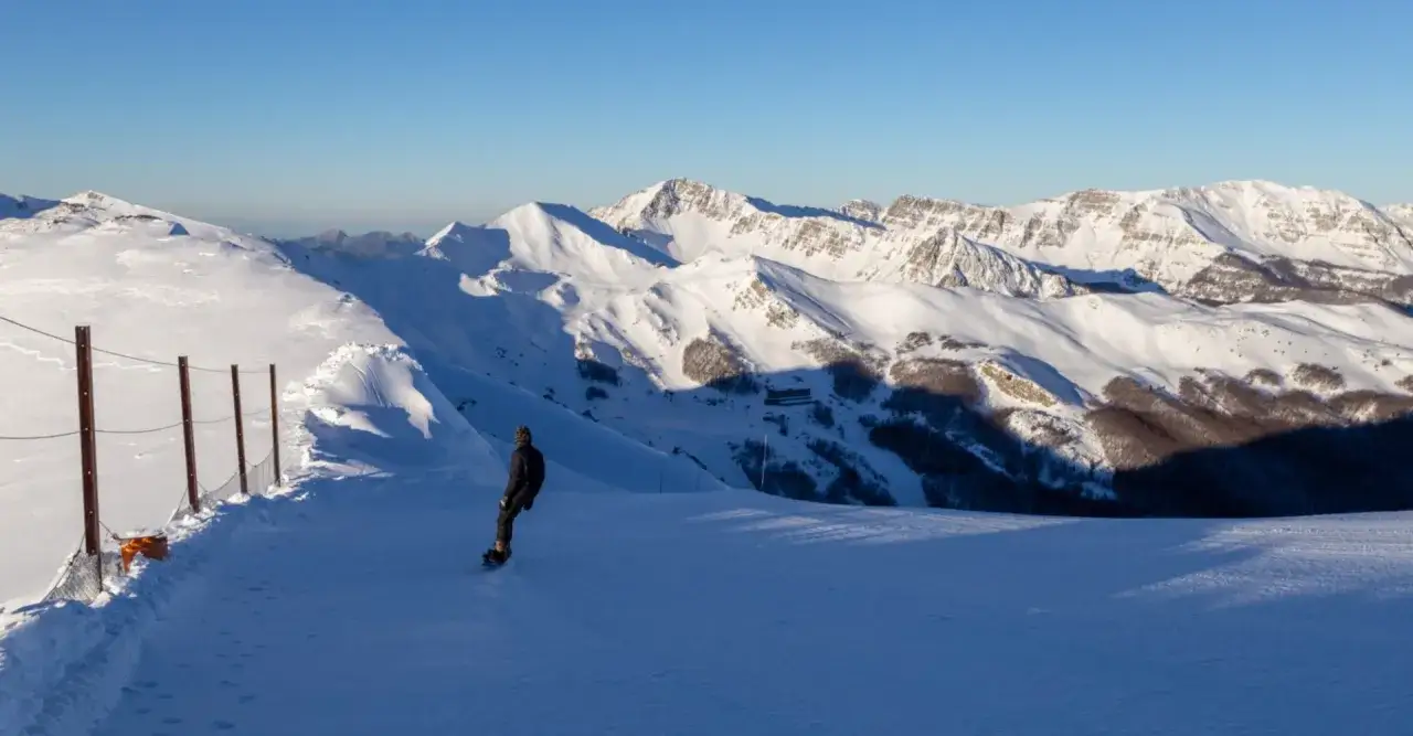Snowboarder scende su una pista innevata, con montagne maestose sullo sfondo. Gli impianti di Abetone offrono panorami mozzafiato.