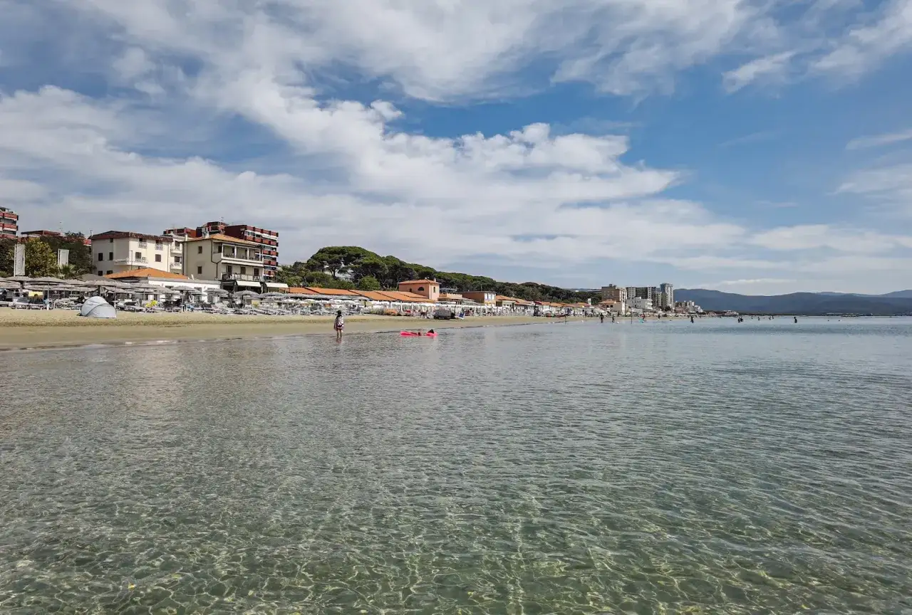 Spiaggia di Follonica con edifici colorati e ombrelloni sulla sabbia. L'acqua cristallina riflette il cielo azzurro con nuvole bianche.