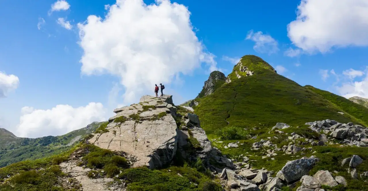 Due escursionisti ammirano il panorama dal passo dell'Abetone, con montagne verdi e cielo azzurro.