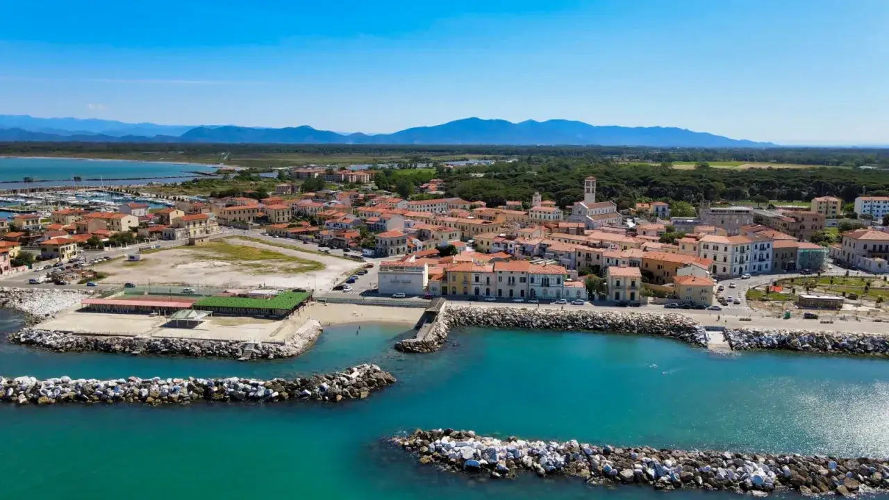 Marina di Pisa: un borgo con case dai tetti rossi, un porto turistico e una spiaggia bagnata da acque cristalline, con montagne sullo sfondo.