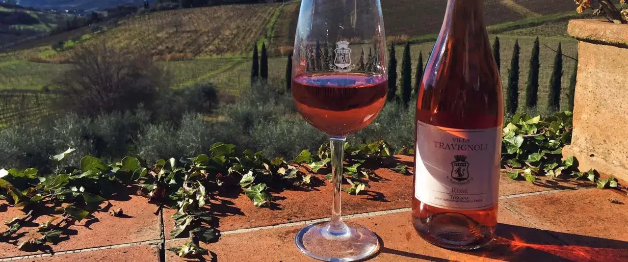 Calice di vino rosato e bottiglia Villa Travignoli, con vigneti toscani sullo sfondo. Un assaggio dei famosi vini bianchi e rosati italiani.