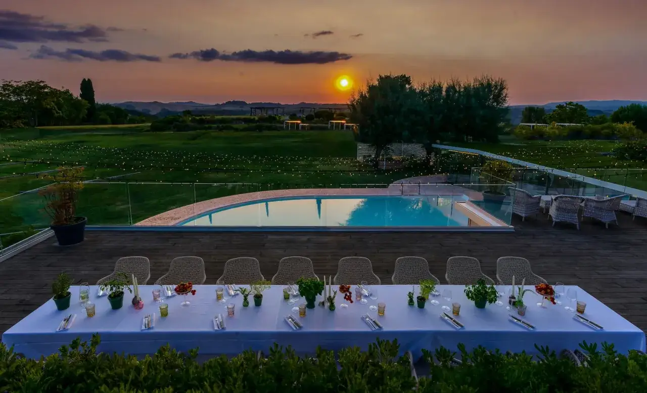 Tavola apparecchiata per una cena all'aperto a Rapolano Terme, con piscina e tramonto sullo sfondo.