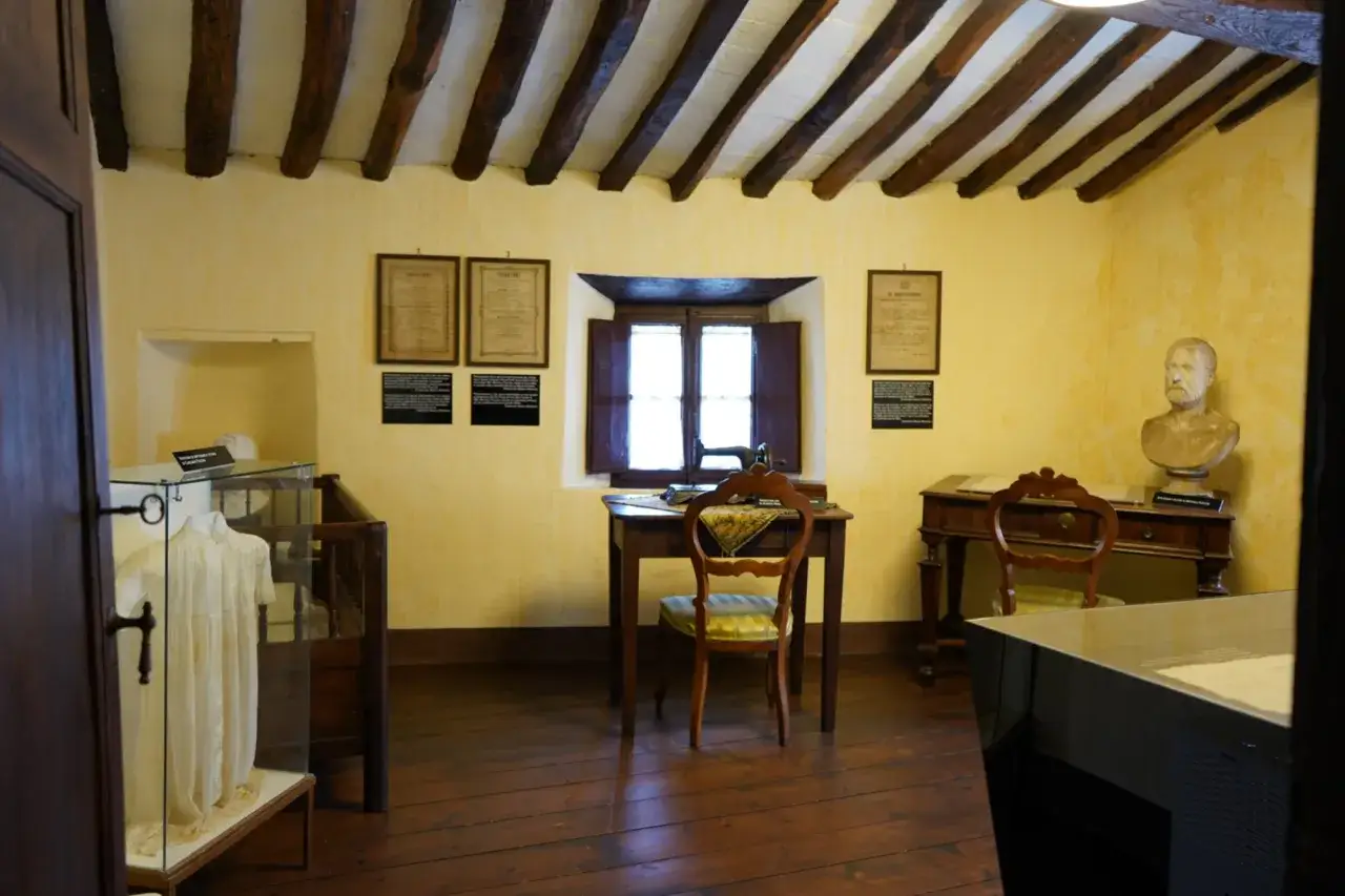 Interno di una casa della memoria: scrivania con macchina da cucire, busto, abito in teca e documenti storici.