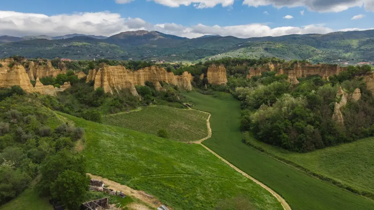 Le suggestive balze del Valdarno, con i loro pinnacoli di terra gialla, si ergono tra verdi colline coltivate e boschi lussureggianti, sotto un cielo azzurro con nuvole bianche.